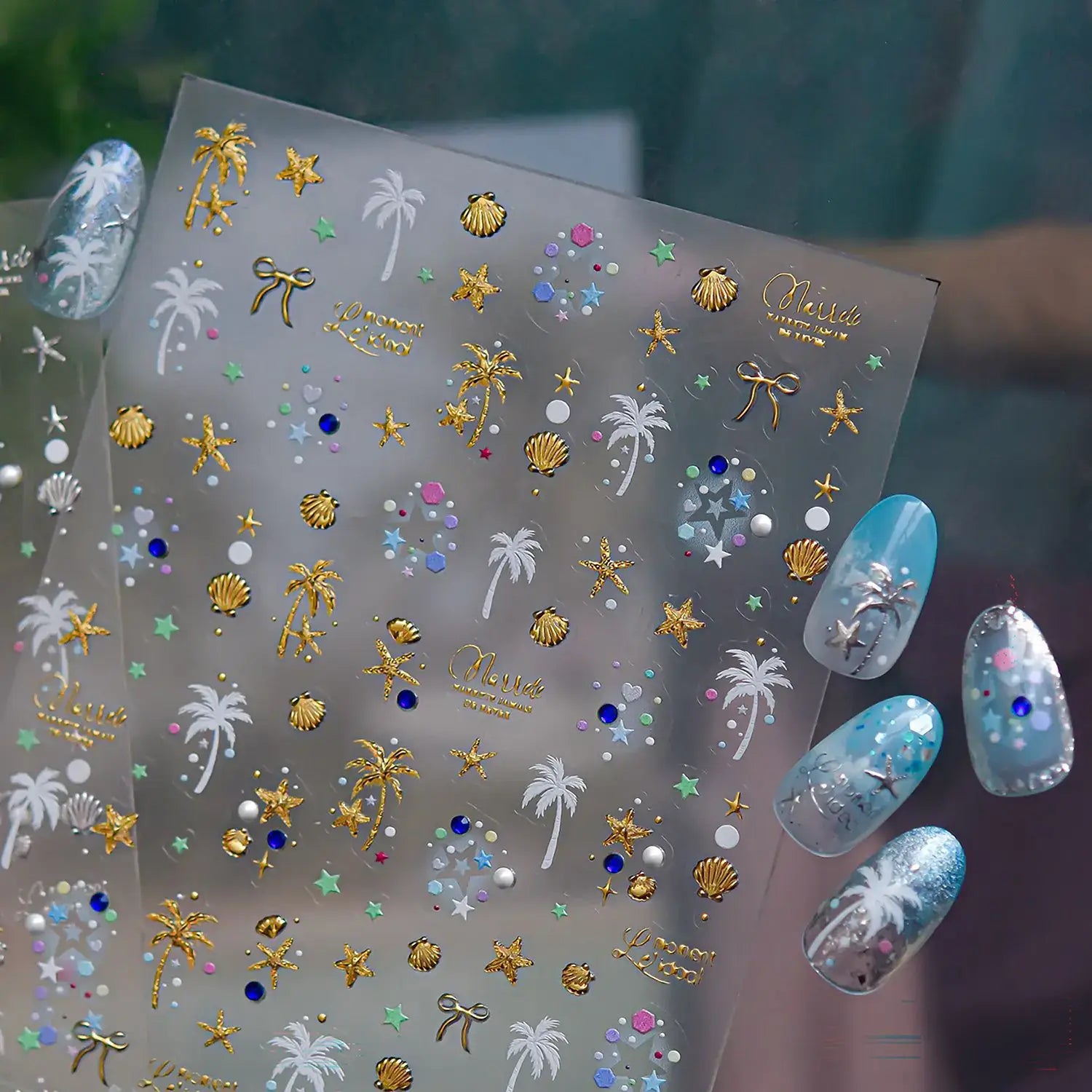 Nail art stickers with palm trees, shells, and stars on a clear sheet, with a hand displaying nails with blue and silver nail art in the background.